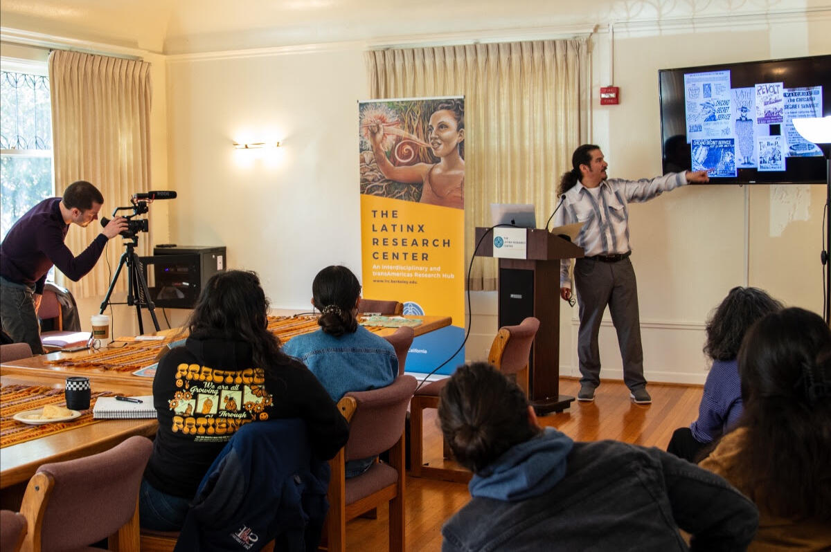 large room with a man giving a lecture poitning to the tv screen with a slideshow another person adjusts a video camera filming the lecture.